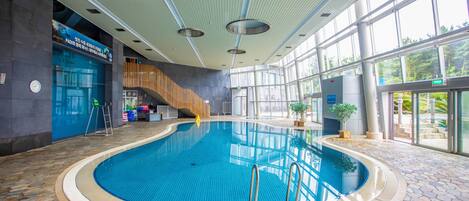 Piscina interior con suelo de baldosas de piedra, escalera de madera y grandes ventanales que dejan entrar luz natural.