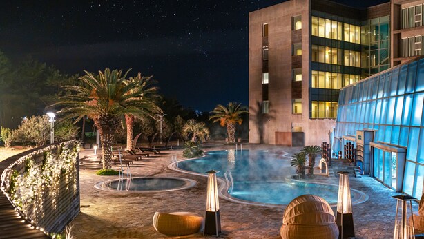 A nighttime resort pool area with multiple pools, lounge chairs, and a modern building with large windows. - Phoenix Island (Seogwipo)