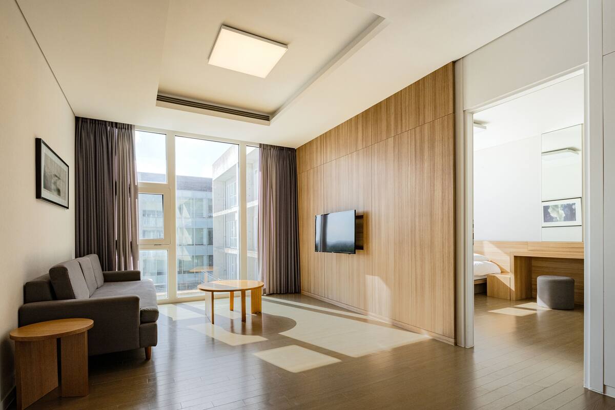 A modern hotel room with a sofa, a small table, a TV mounted on the wall, and a large window with curtains.