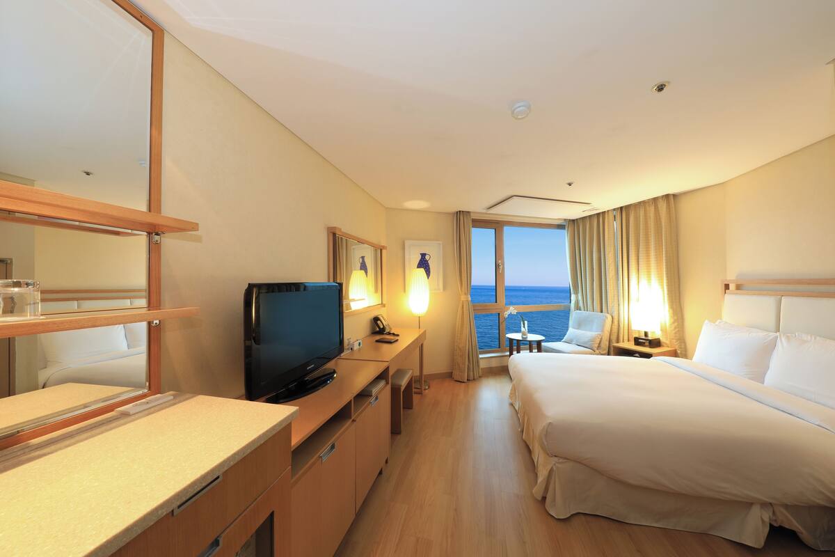 Une chambre d’hôtel avec un lit, un bureau avec une télévision, une chaise, une petite table et une fenêtre donnant sur la mer.