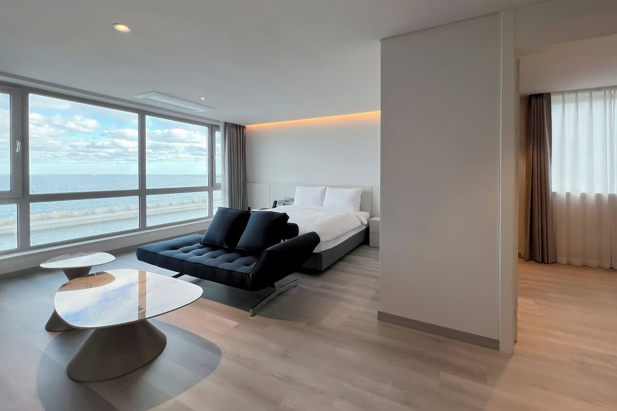 Une chambre d’hôtel moderne dotée d’une grande fenêtre donnant sur la mer, d’un lit, d’un canapé et d’une petite table ronde.
