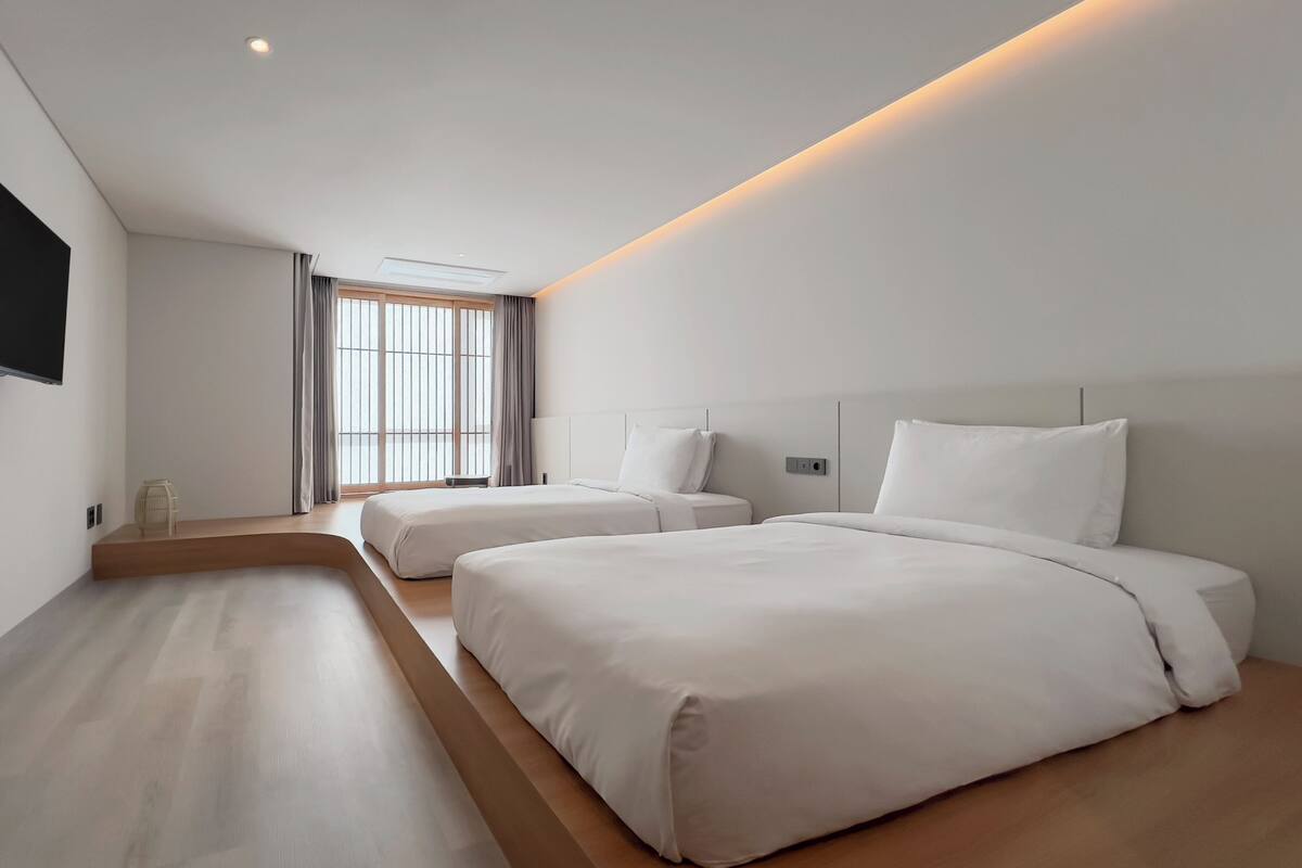 Une chambre d’hôtel moderne avec deux lits, un banc en bois, un téléviseur à écran plat et une grande fenêtre avec des rideaux.