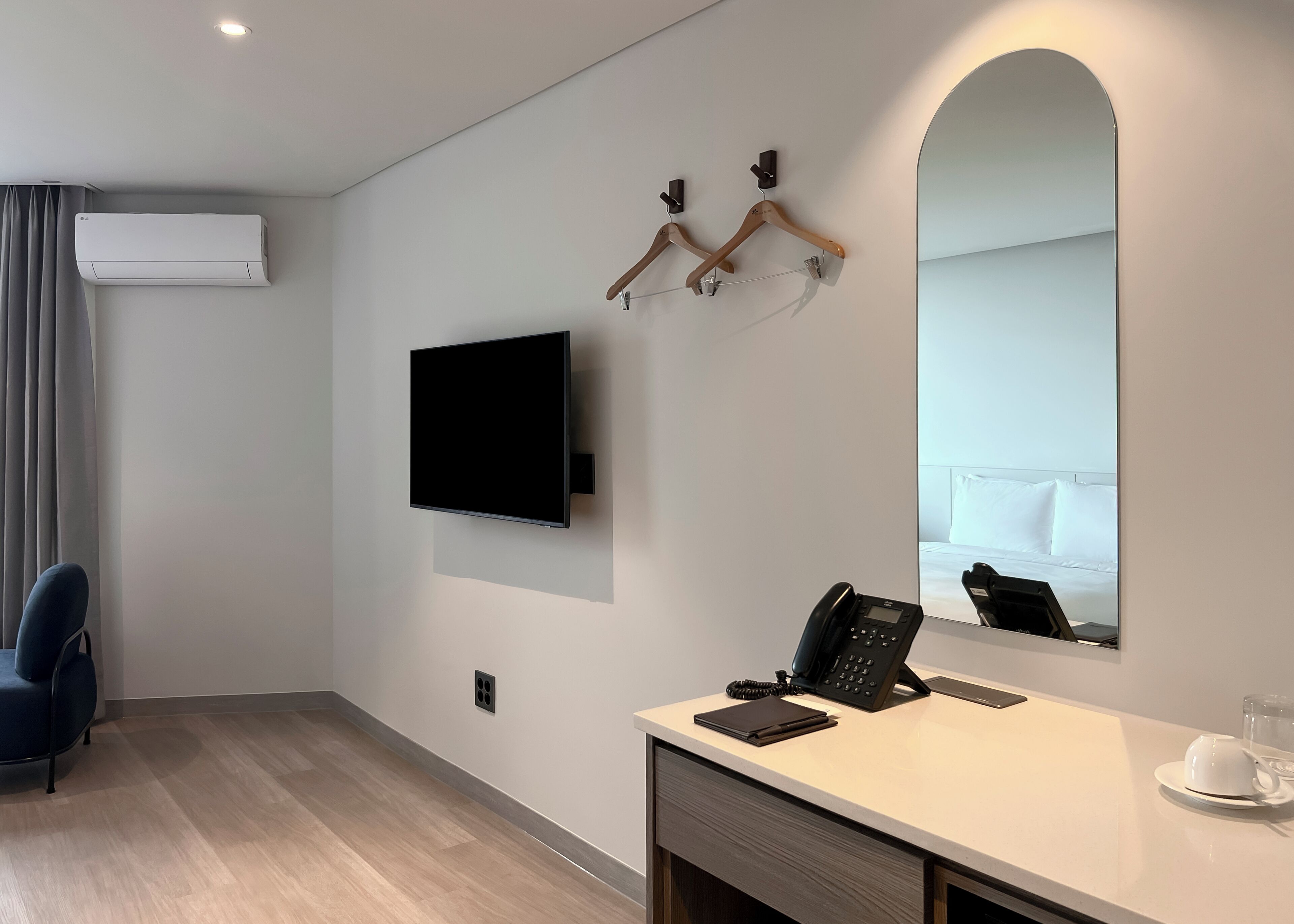 Quarto de hotel moderno com televisão de tela plana na parede, uma mesa com telefone e um espelho refletindo o ambiente.