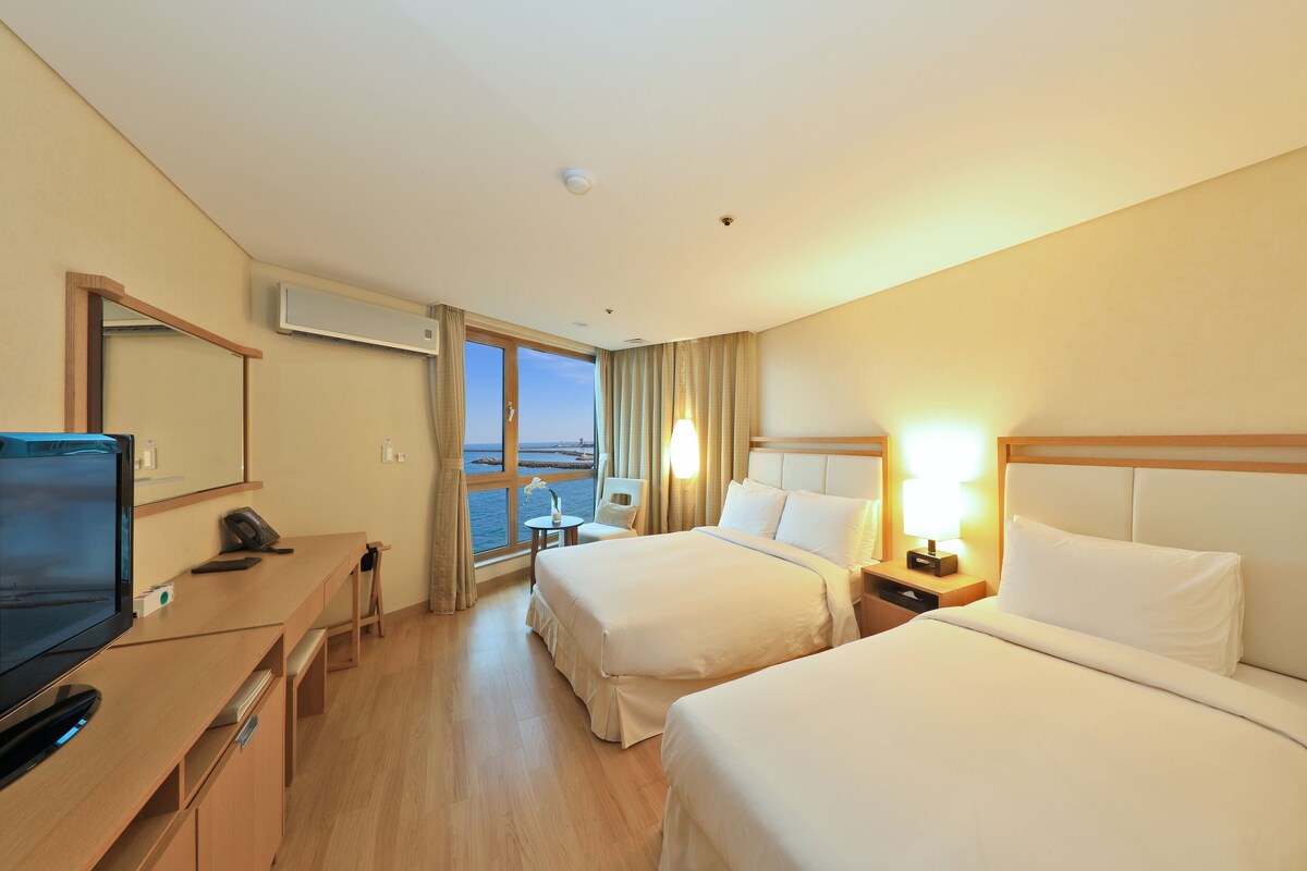 Une chambre d’hôtel avec deux lits, un bureau avec une télévision, une chaise et une fenêtre donnant sur la mer.