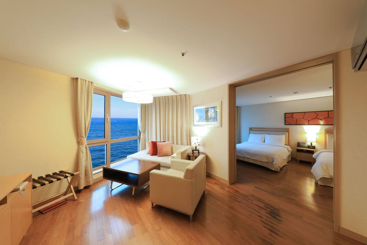 Une chambre d’hôtel avec un plancher en bois, un bureau avec une chaise, un canapé, un lit et une vue sur la mer par une grande fenêtre.