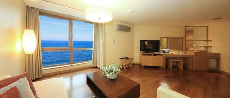 Habitación de hotel con un amplio ventanal que ofrece vistas al mar, un sofá, una mesa de centro, un televisor y una kitchenette.