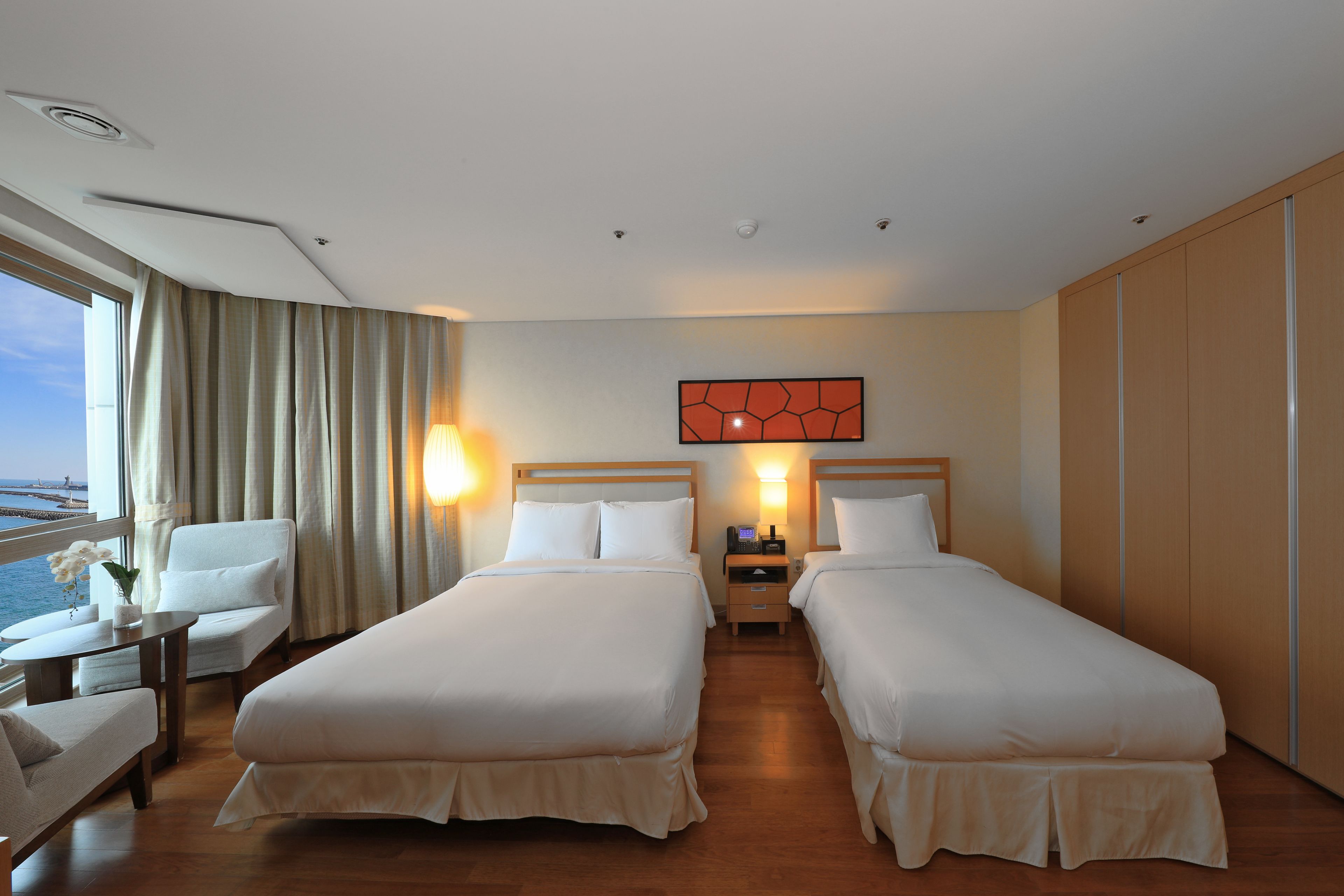 A hotel room with two beds, a sitting area with a chair and table, a view of the sea, and a decorative wall piece.