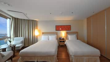 A hotel room with two beds, a sitting area with a chair and table, a view of the sea, and a decorative wall piece.