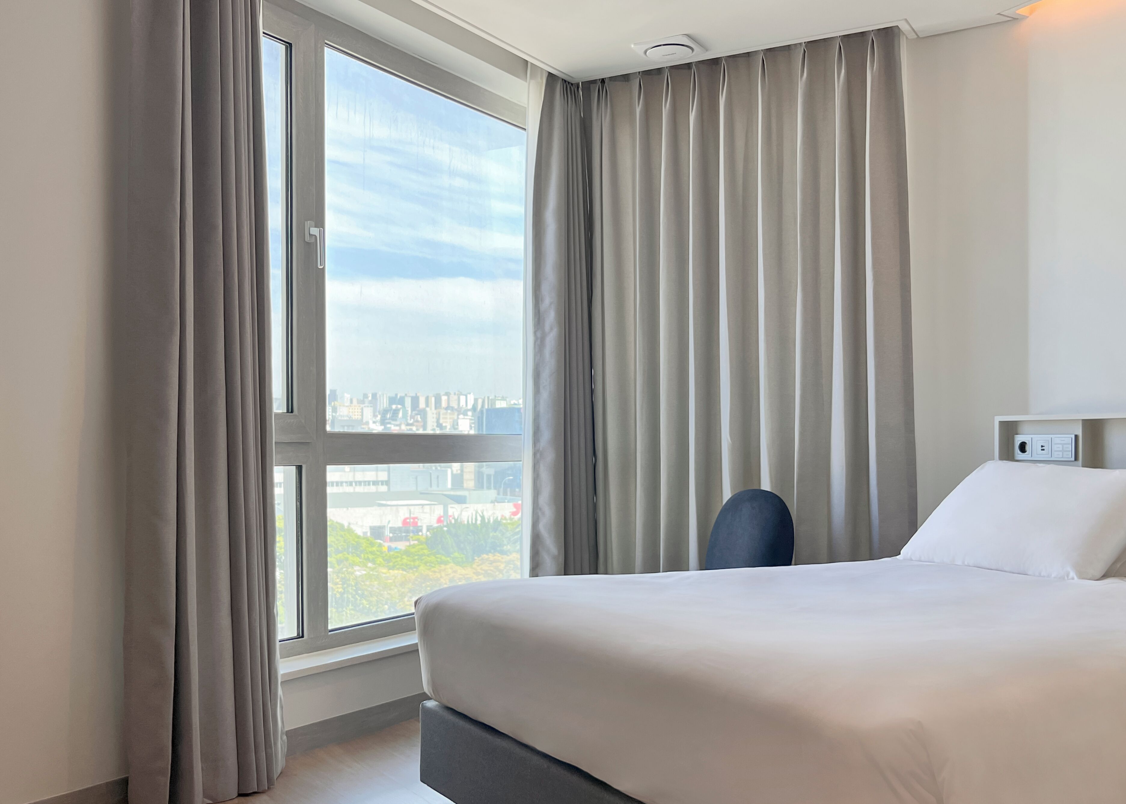 A hotel room with a large window, a bed, a chair, and a control panel on the wall.