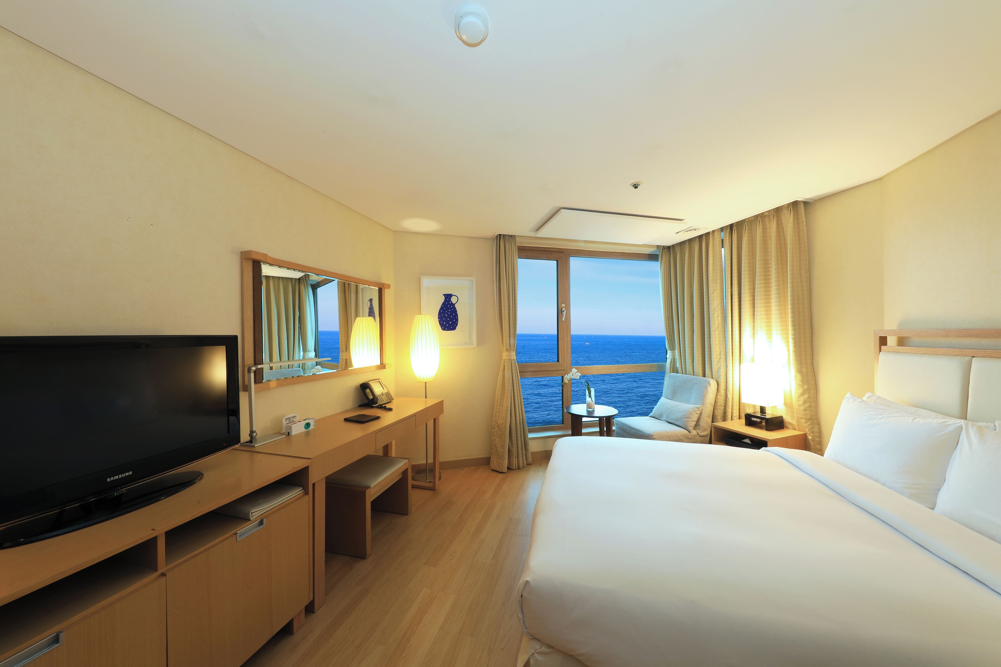 Ein Hotelzimmer mit einem großen Bett, einem Schreibtisch mit Flachbildfernseher, einem Sessel, einem kleinen Tisch und Meerblick durch das Fenster.