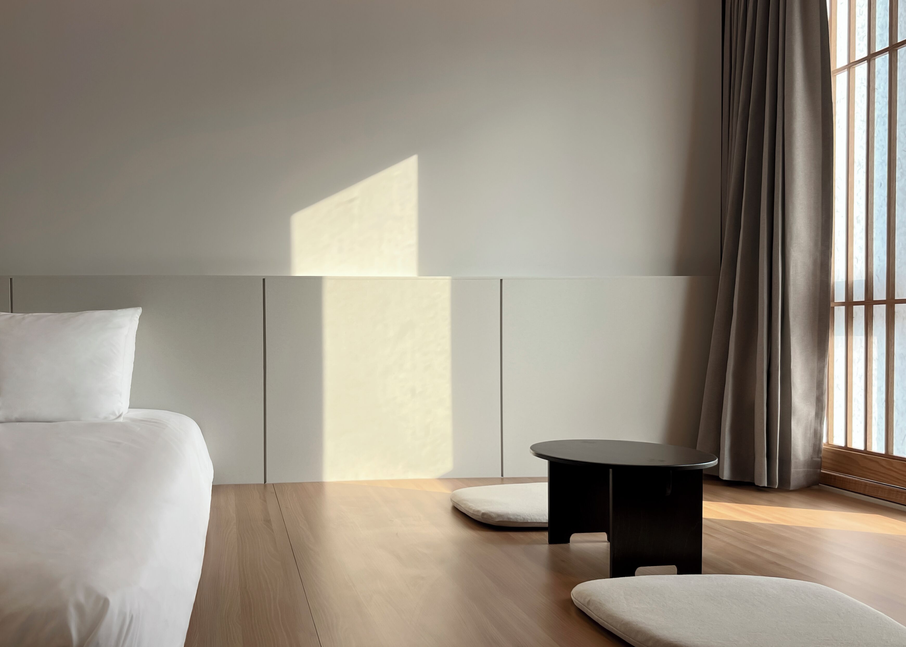 A minimalist hotel room with a bed, a small table, and a window with curtains.