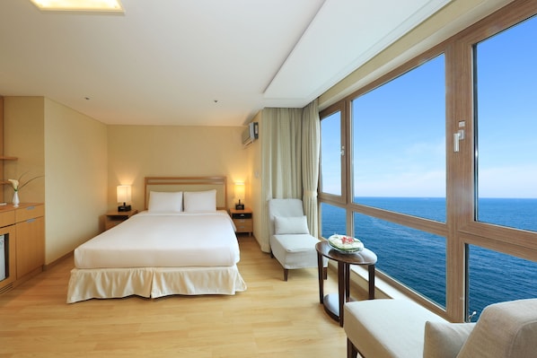 A hotel room with a large bed, a chair, a small table with a flower, and a view of the ocean.