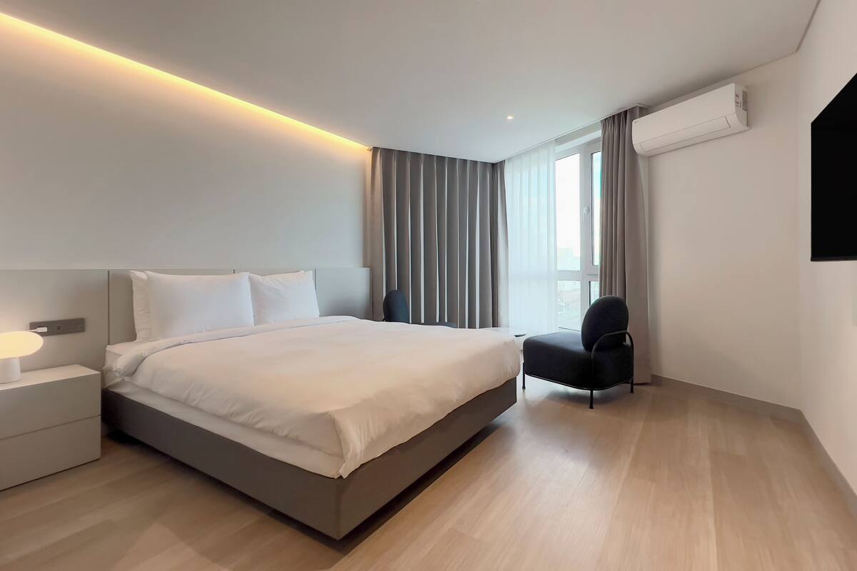 Une chambre d’hôtel moderne avec un grand lit, une table de chevet avec une lampe, une chaise et une grande fenêtre avec des rideaux.