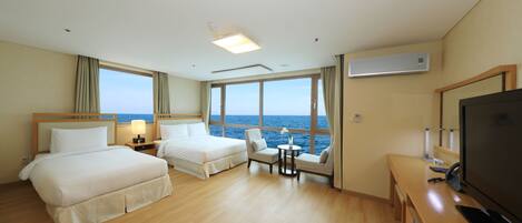 A hotel room with two beds, a TV, a desk, and a view of the sea.