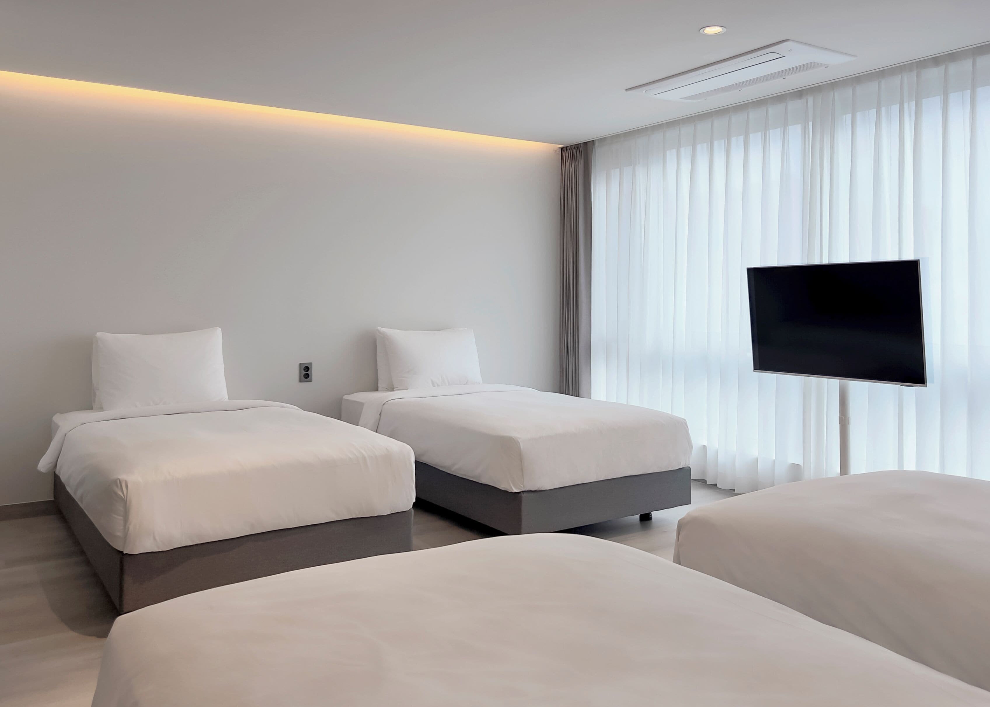 Una habitación de hotel moderna con dos camas individuales, un televisor de pantalla plana montado en la pared y un ventanal con cortinas translúcidas.