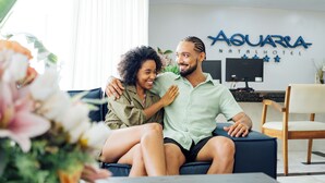 A couple sitting on a couch in a hotel lobby with a reception desk and a sign that reads 'AQUARIA NATALHOTEL'. - Aquaria Natal Hotel (Natal)