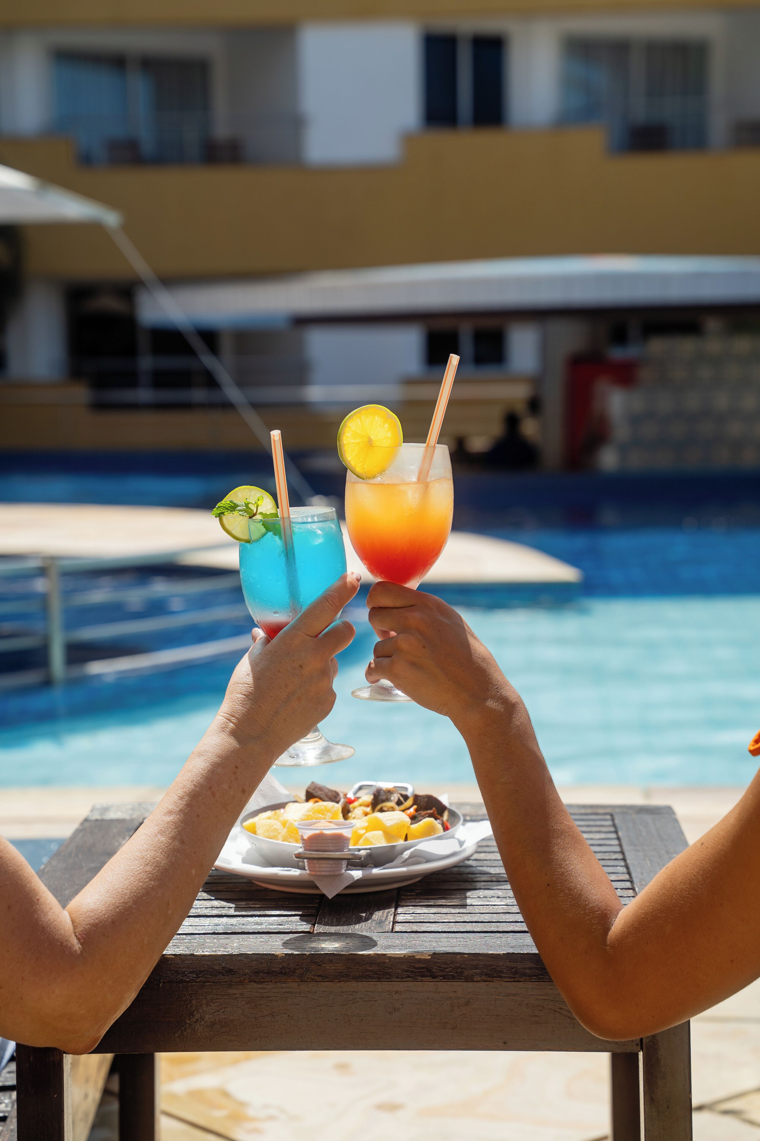 Dos personas brindando con bebidas en una mesa junto a la piscina, con una piscina y un edificio al fondo.