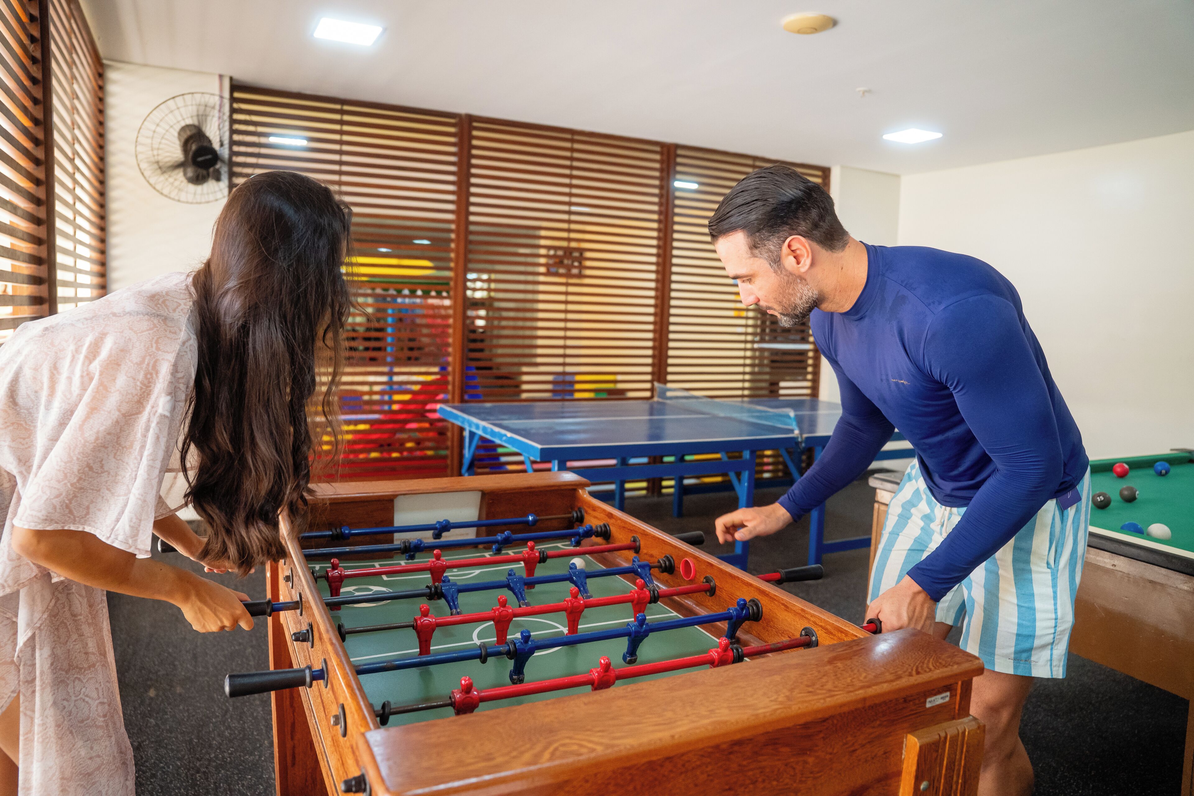 A foosball table, a pool table, and a fan in a room with wooden blinds.