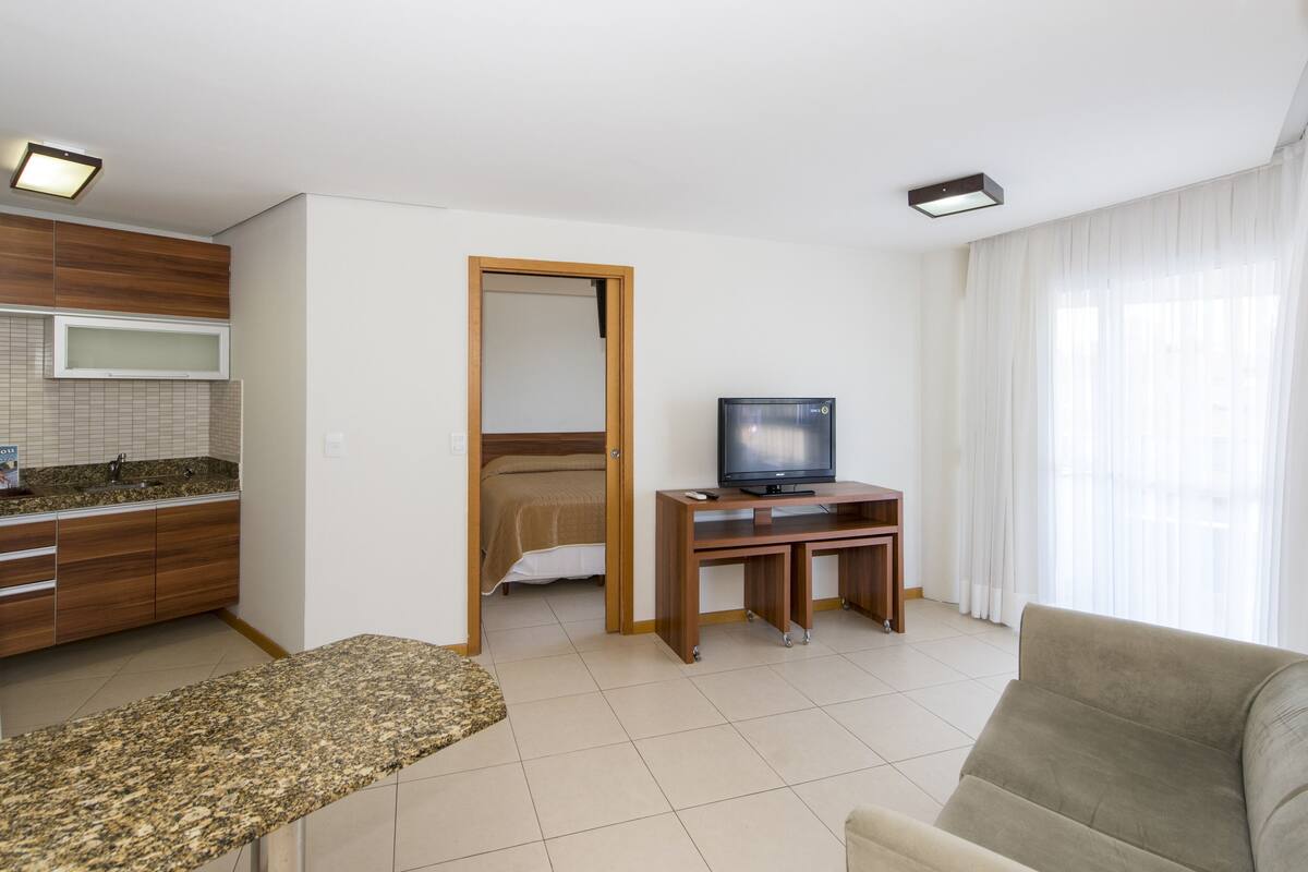 A modern living room with a television, a sofa, a coffee table, and a kitchen area with a microwave and sink.