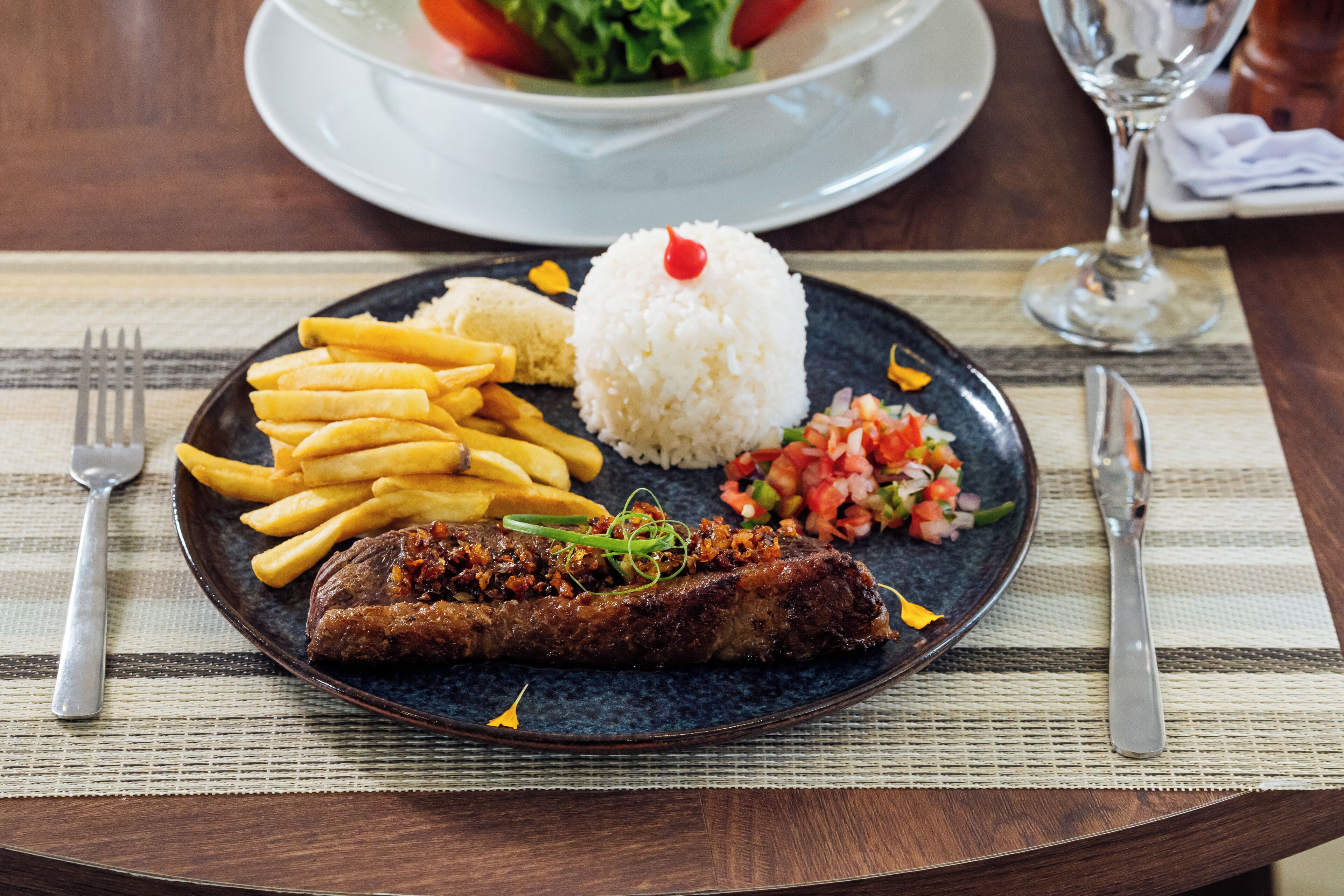 A plate with a meat dish, fries, rice, and a side salad on a table with a placemat.
