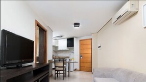 A living room with a flat-screen TV on a dark wooden stand, a grey sofa, and a kitchen area with a wooden door and white cabinets. - Aquaria Natal Hotel (Natal)
