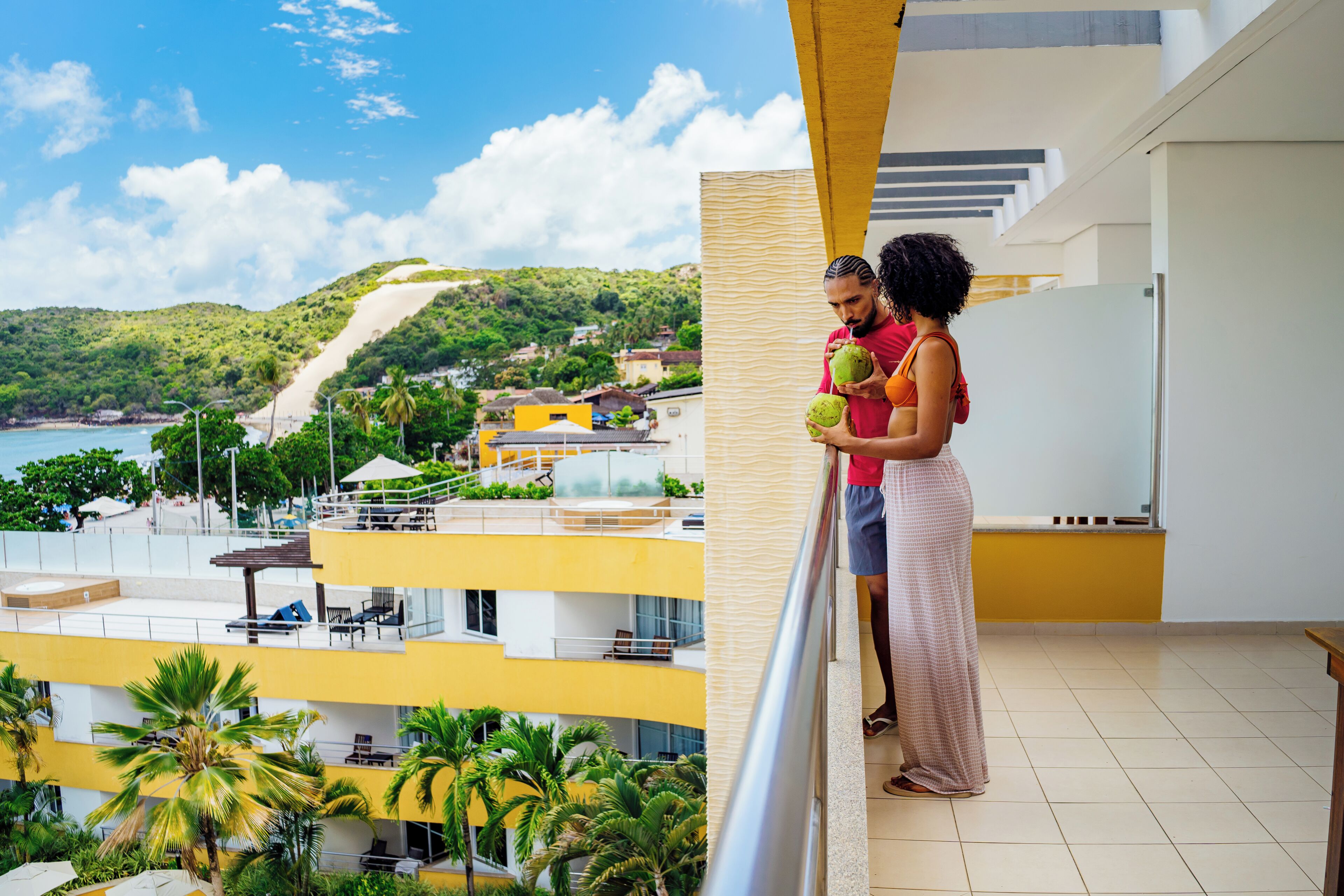 Un balcón con vista a una playa y colinas, un edificio amarillo y una persona sosteniendo un coco.