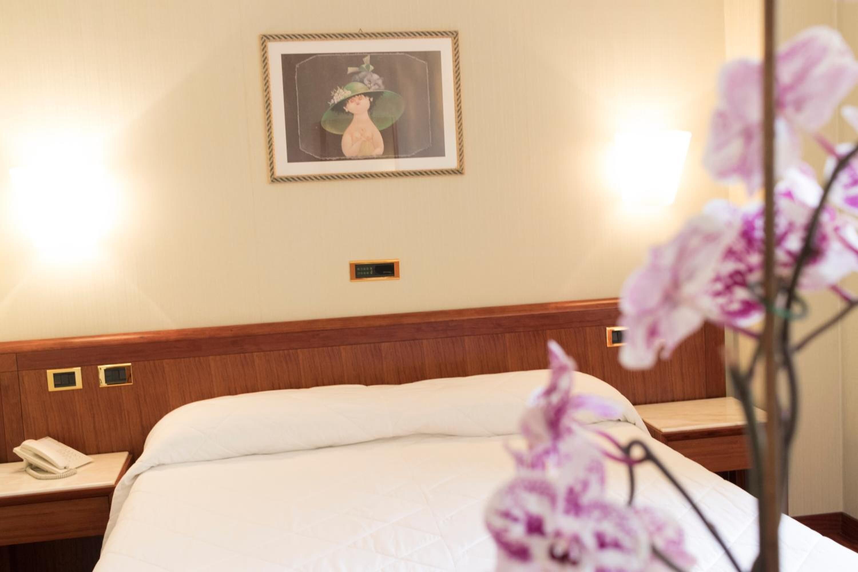 Habitación de hotel con una cama, una mesita de noche con un teléfono, un cuadro en la pared y un jarrón con flores.