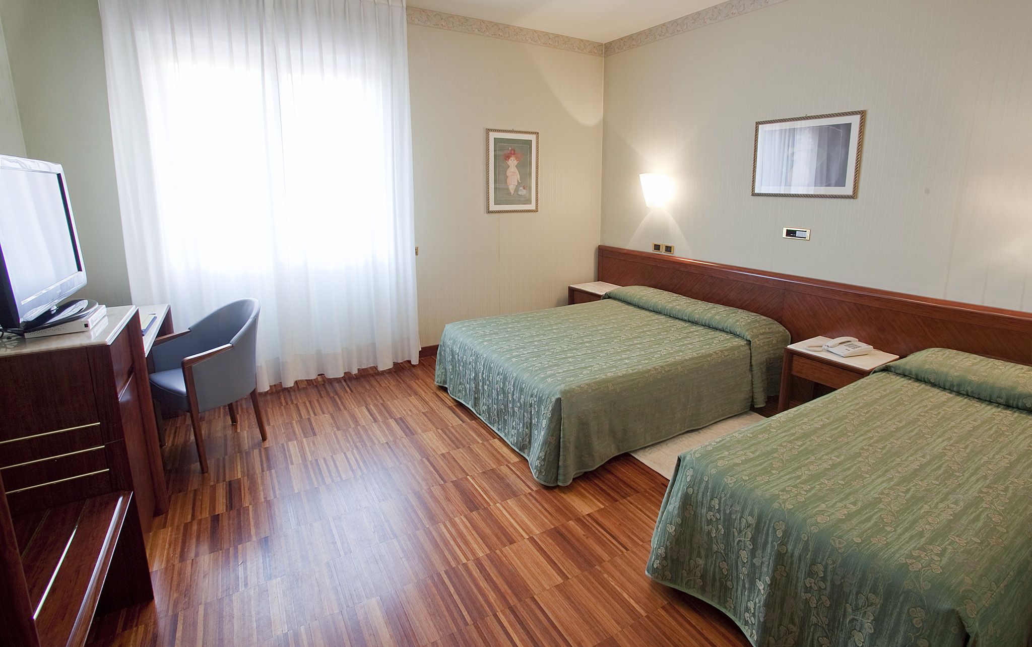 A hotel room with two single beds, a desk with a computer, a chair, and a window with curtains.