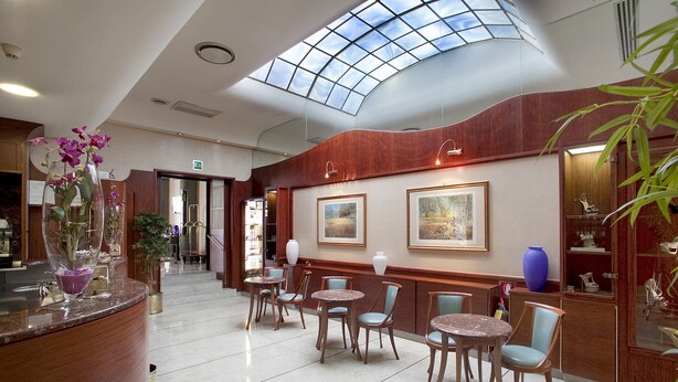A hotel lobby with a high ceiling, a reception desk, several tables and chairs, framed artwork on the walls, and a large skylight. - Palace Hotel (Civitanova Marche)