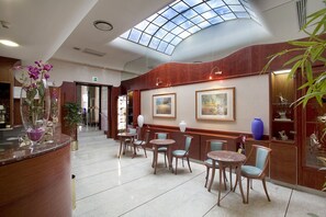 Een hotel lobby met een hoge plafond, een receptiebalie, diverse tafels en stoelen, kunstwerken in lijsten aan de muren, en een groot dakvenster.