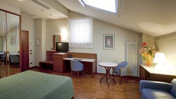 A hotel room with a bed, a desk, a chair, a small table, a lamp, and a radiator.