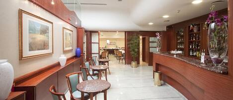 A hotel lobby with wooden paneling, a bar area with a marble countertop, and a dining area with round tables and chairs.