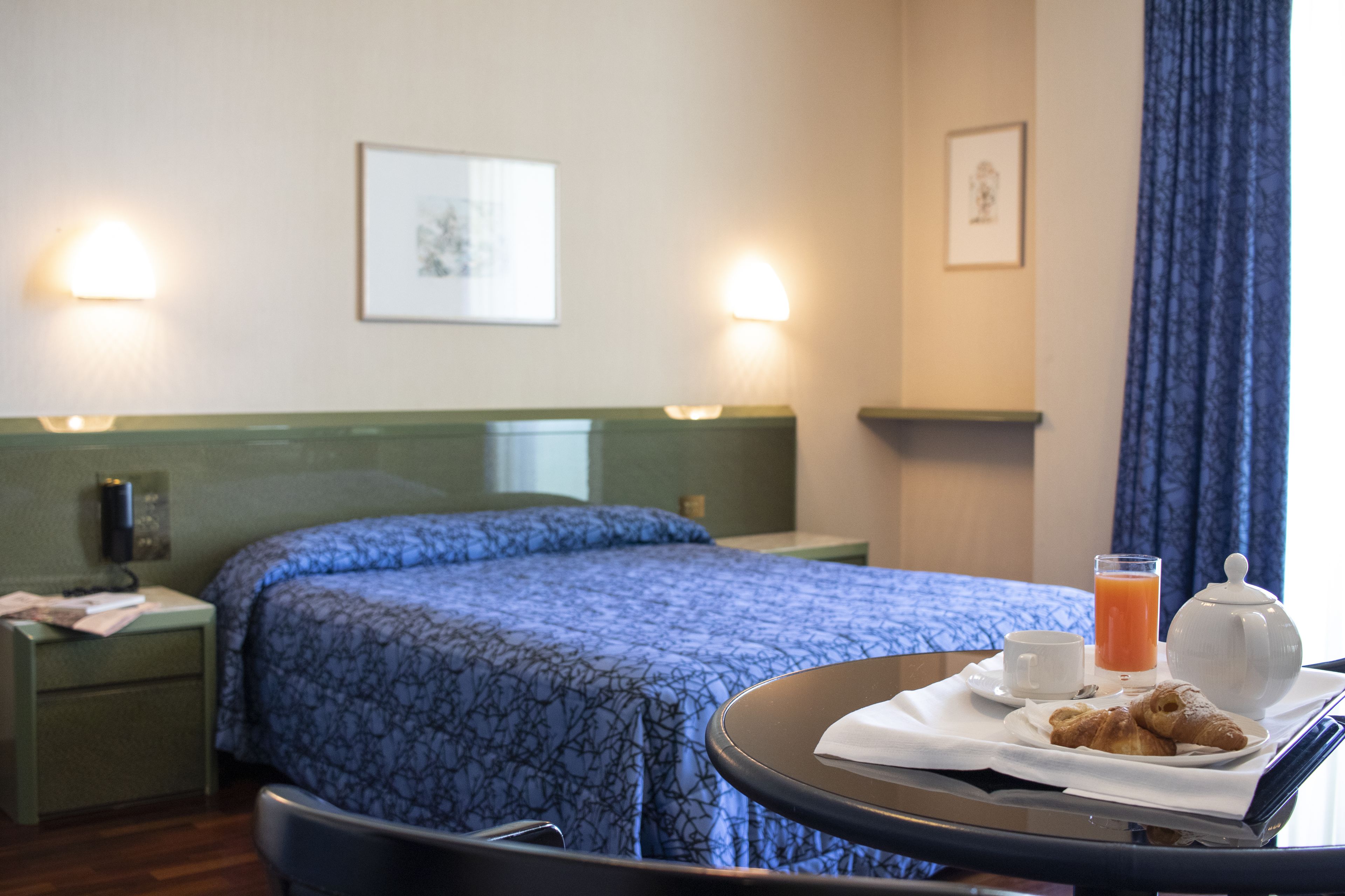 Ein Hotelzimmer mit einem ordentlich bezogenen Bett, einem Nachttisch mit Lampe sowie einem Tablett mit Croissants, einer Tasse Kaffee und einem Glas Orangensaft.