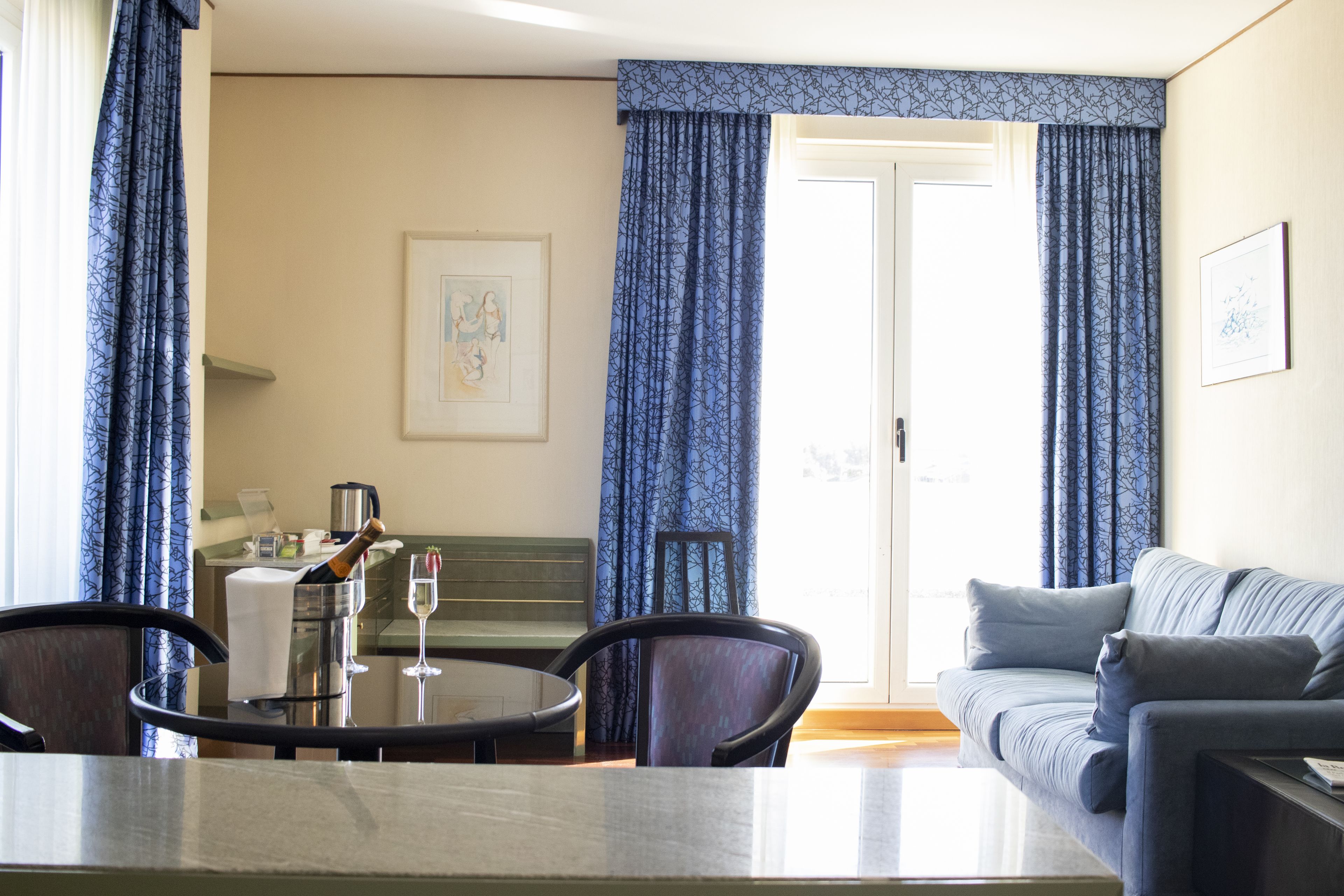 A hotel room with a dining area, a sofa, and a large window with curtains.