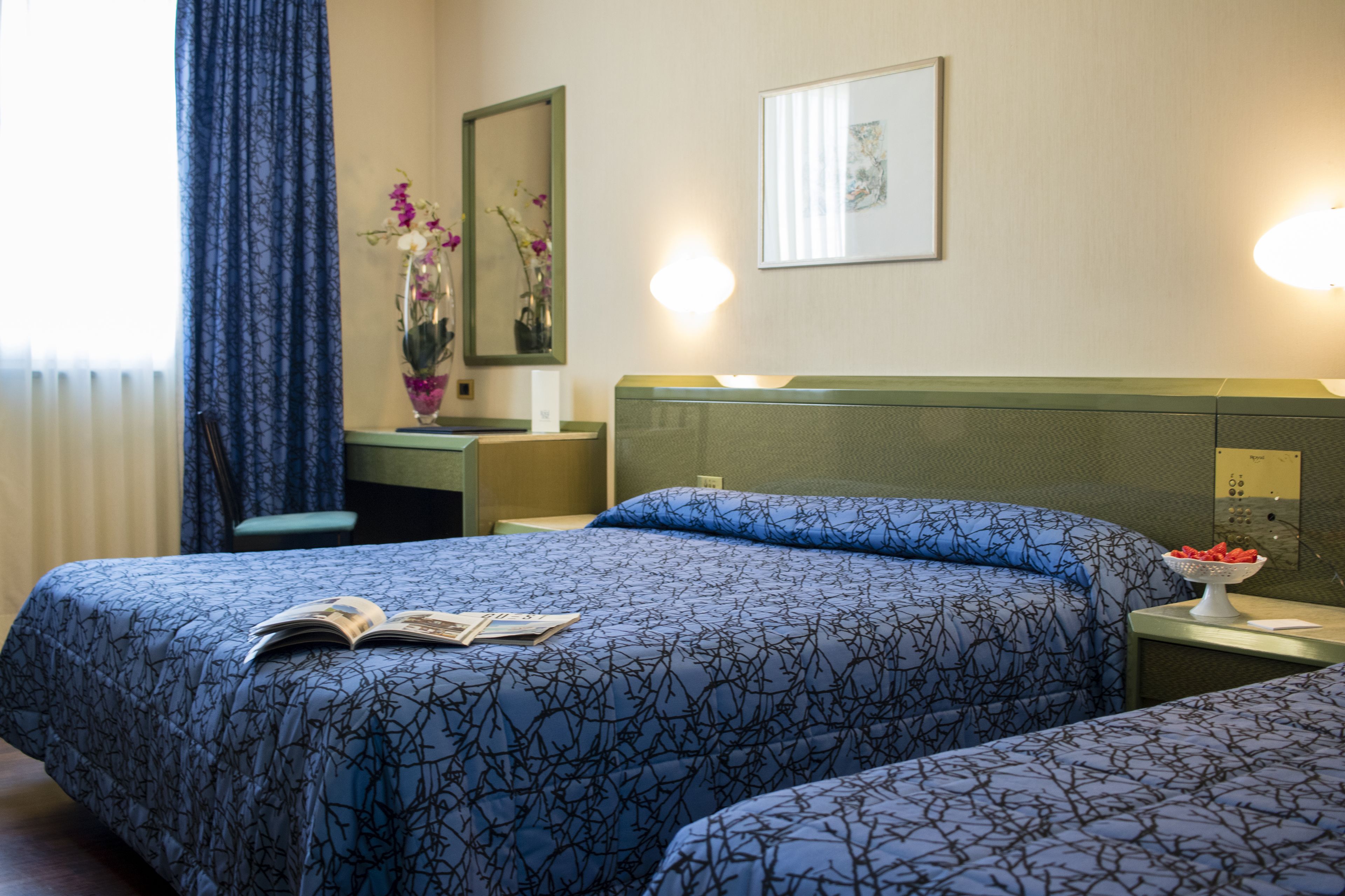 Une chambre d’hôtel avec deux lits, un bureau, une chaise, un vase avec des fleurs, un tableau encadré et une table de chevet avec une lampe.