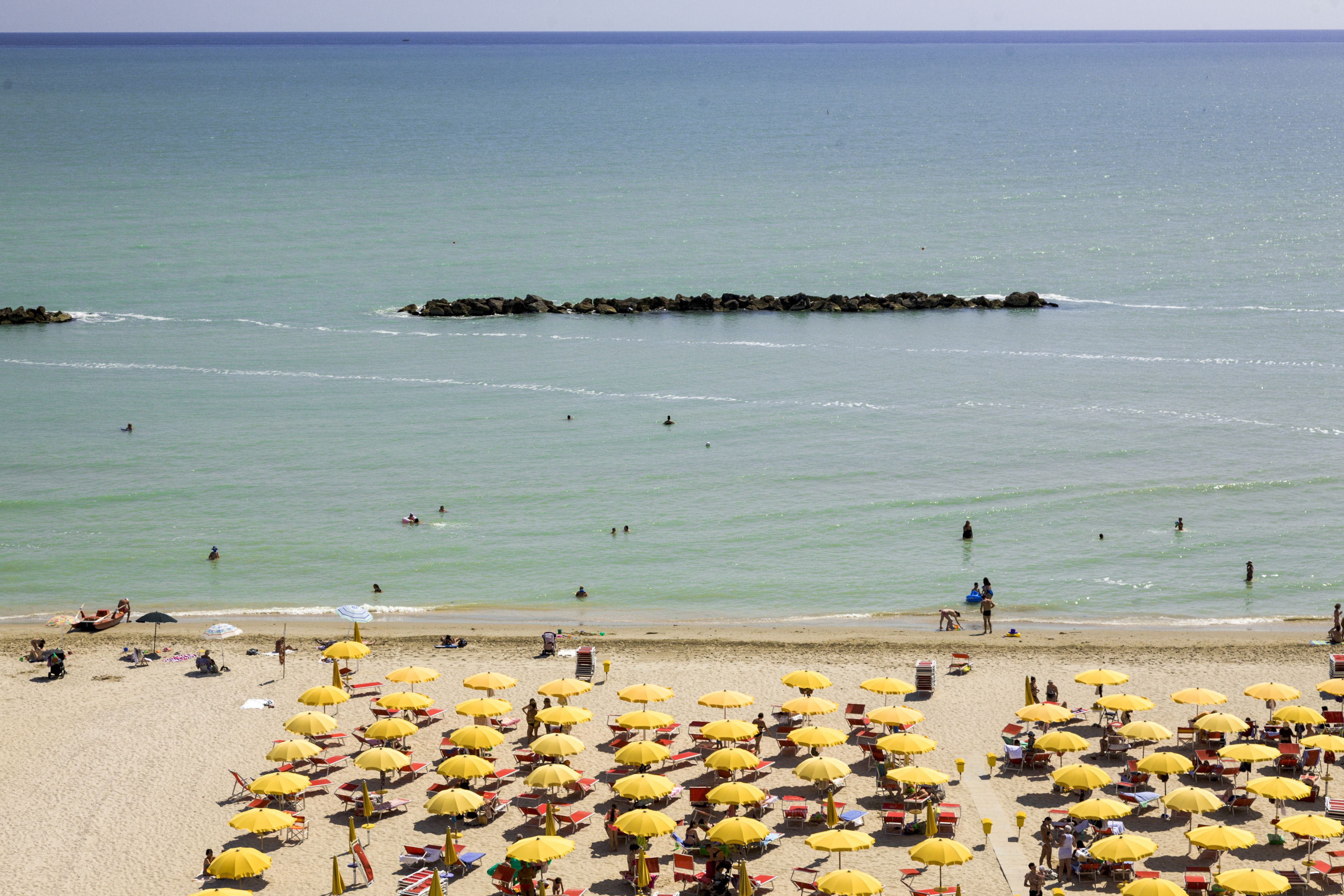 A beach with numerous yellow umbrellas and sun loungers, a rocky breakwater in the water, and people swimming and relaxing on the shore.