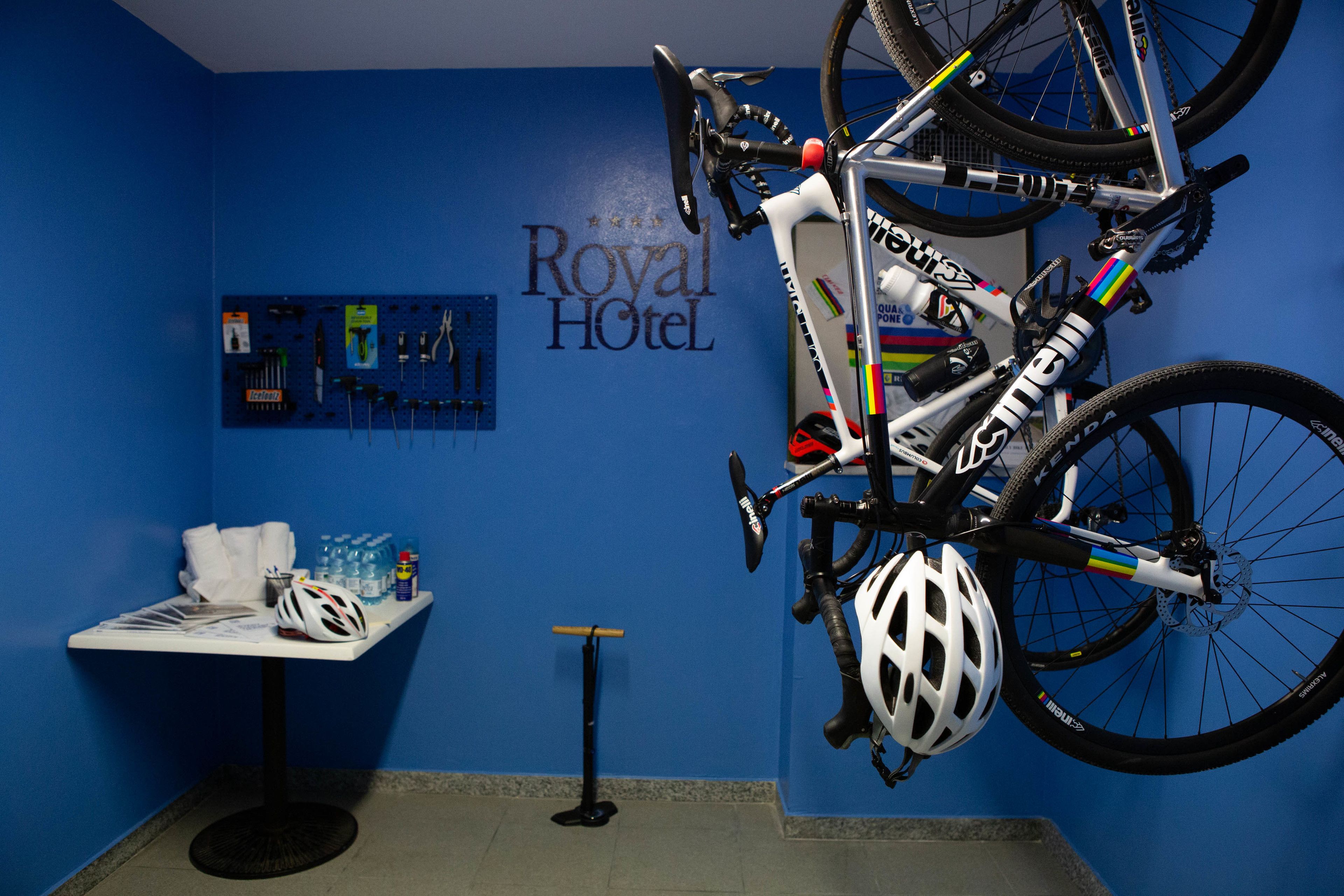A bicycle hanging on the wall with a helmet beside it, a table with water bottles and a helmet, and a wall with the text "Royal Hotel".