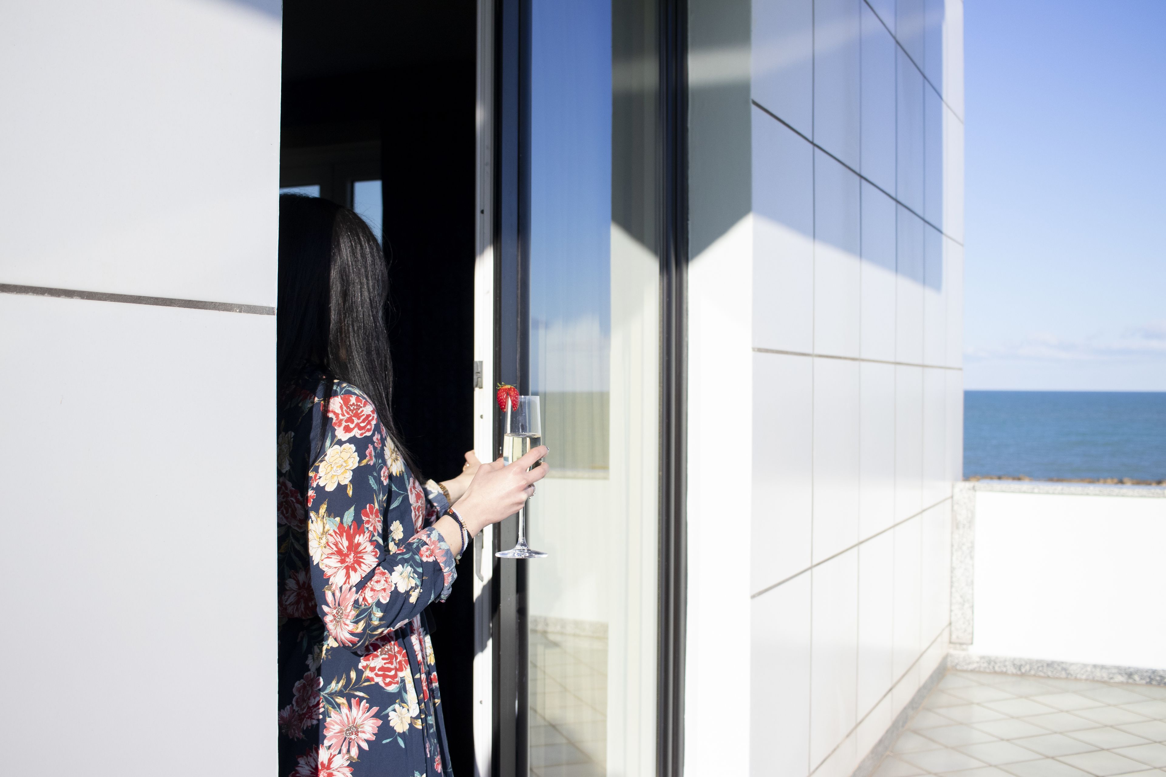 A person in a floral dress holding a glass, standing at a glass door with a view of the sea.