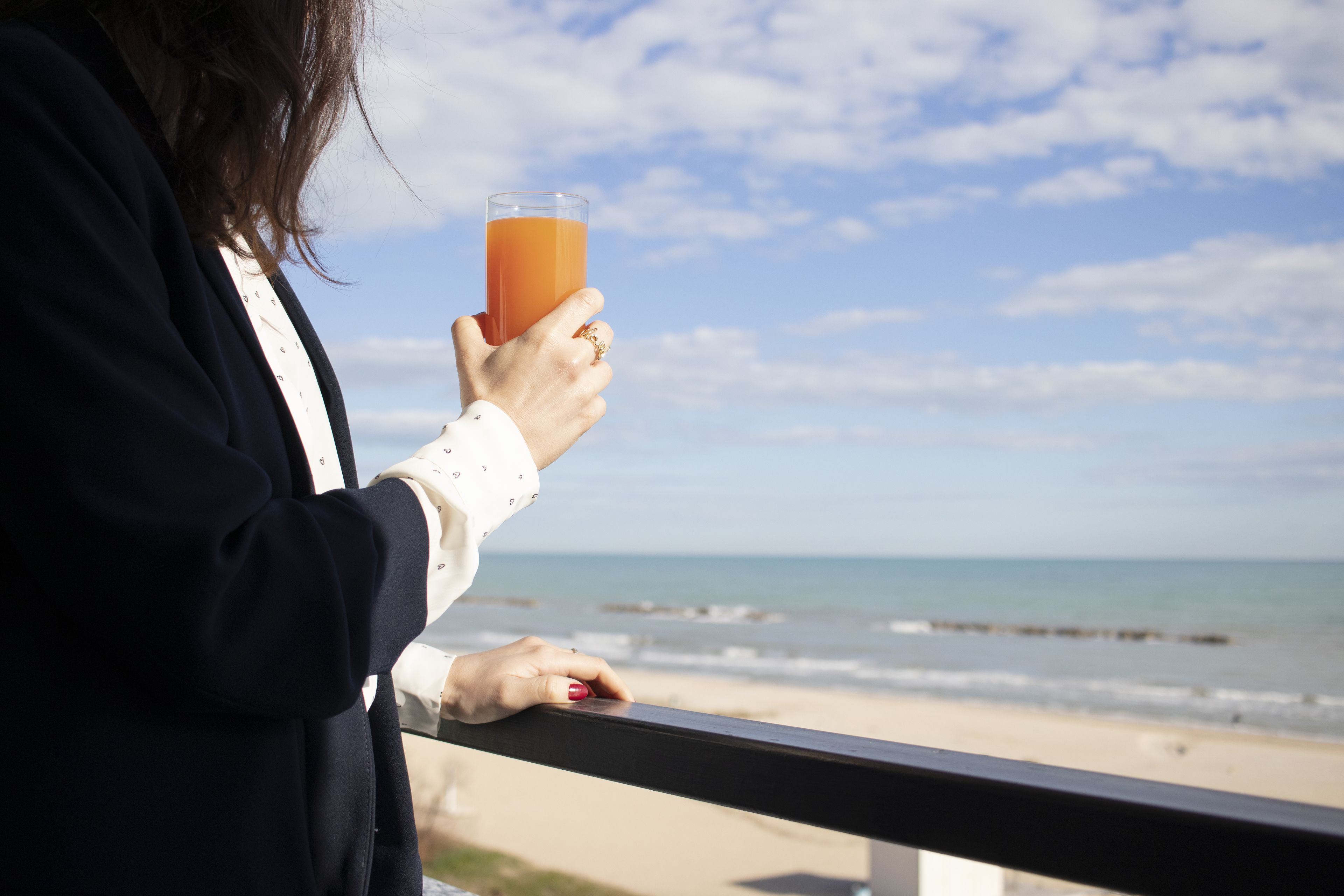 Una persona sostiene un vaso de jugo de naranja, contemplando una playa con olas y un cielo despejado.