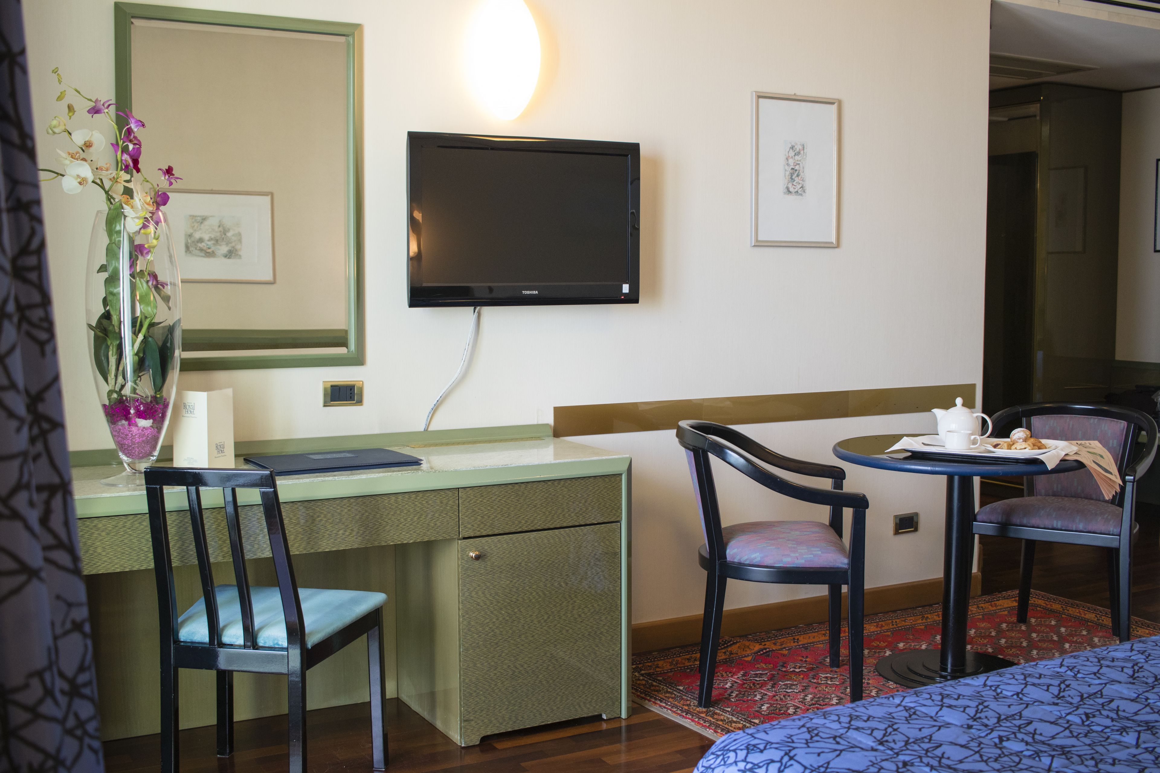 Habitación de hotel con escritorio, silla, televisor en la pared y cama con colcha estampada.