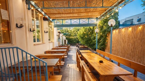 An outdoor dining area with wooden benches and tables, a covered walkway, and a view of greenery.