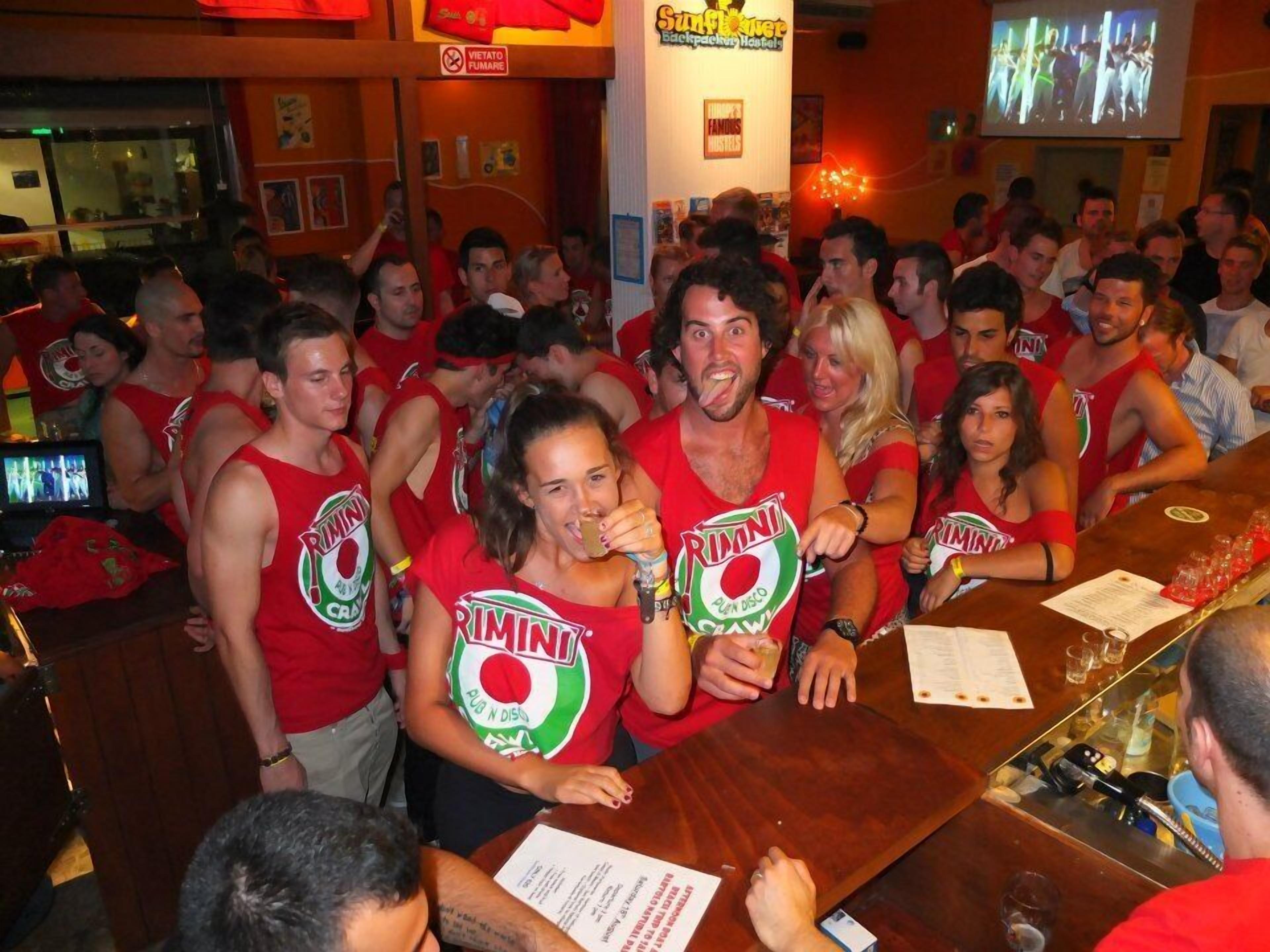 Un bar concurrido con gente usando camisetas rojas de 'RIMINI', una pantalla de televisión y un cartel que dice 'Vietato Fumare'.