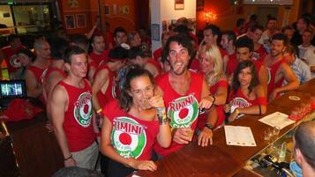 Un bar concurrido con gente usando camisetas rojas de 'RIMINI', una pantalla de televisión y un cartel que dice 'Vietato Fumare'.