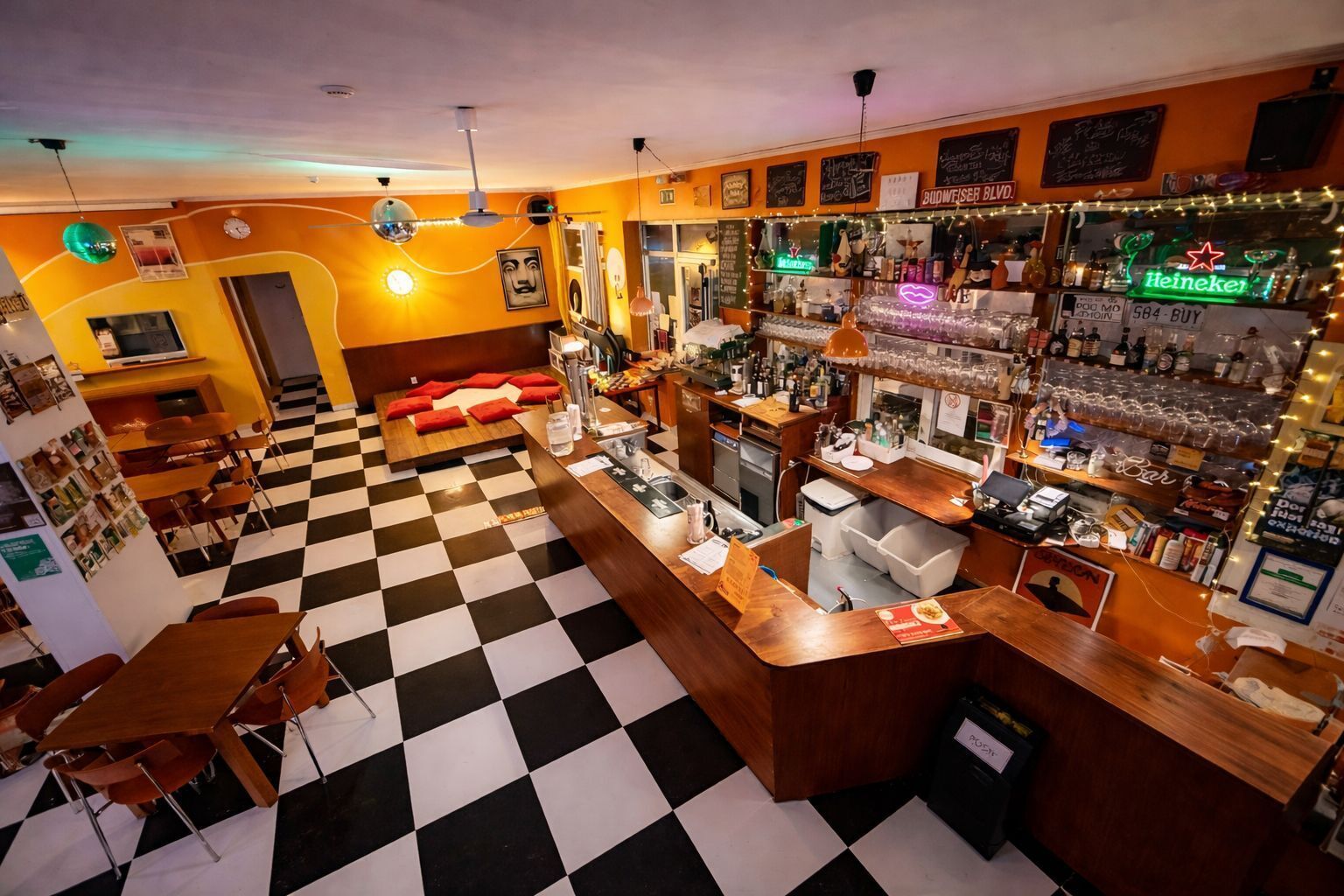 Un espace bar avec un sol à carreaux, un comptoir orné d'un néon, et diverses bouteilles et verres disposés sur des étagères.
