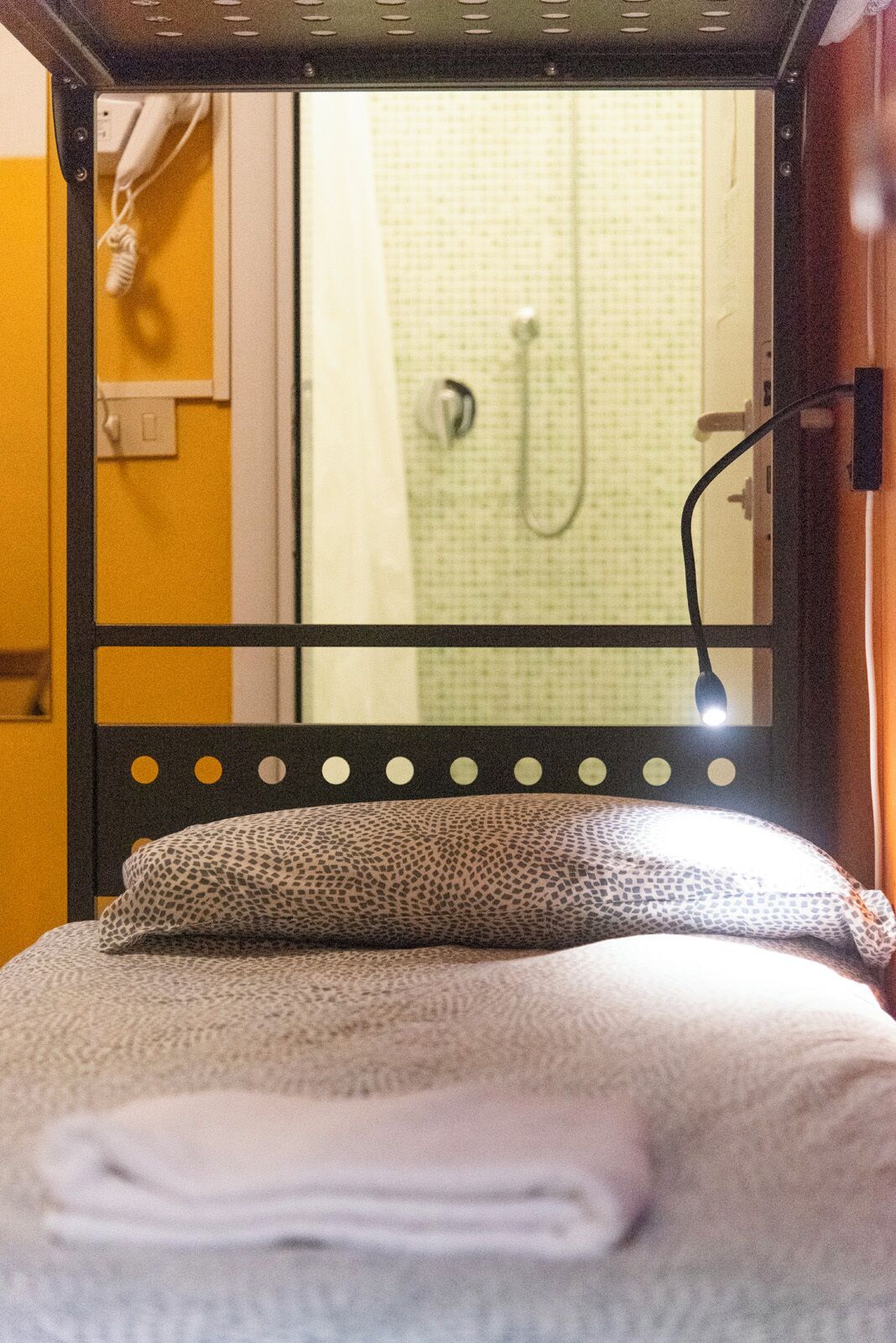 Uma cama com travesseiro estampado e toalha dobrada, cabeceira preta com recortes circulares, e área de chuveiro visível com chuveiro e painel de controle.