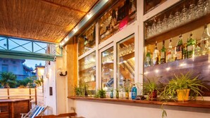 An outdoor bar area with wooden tables and chairs, a bar counter, and shelves with bottles and glasses.