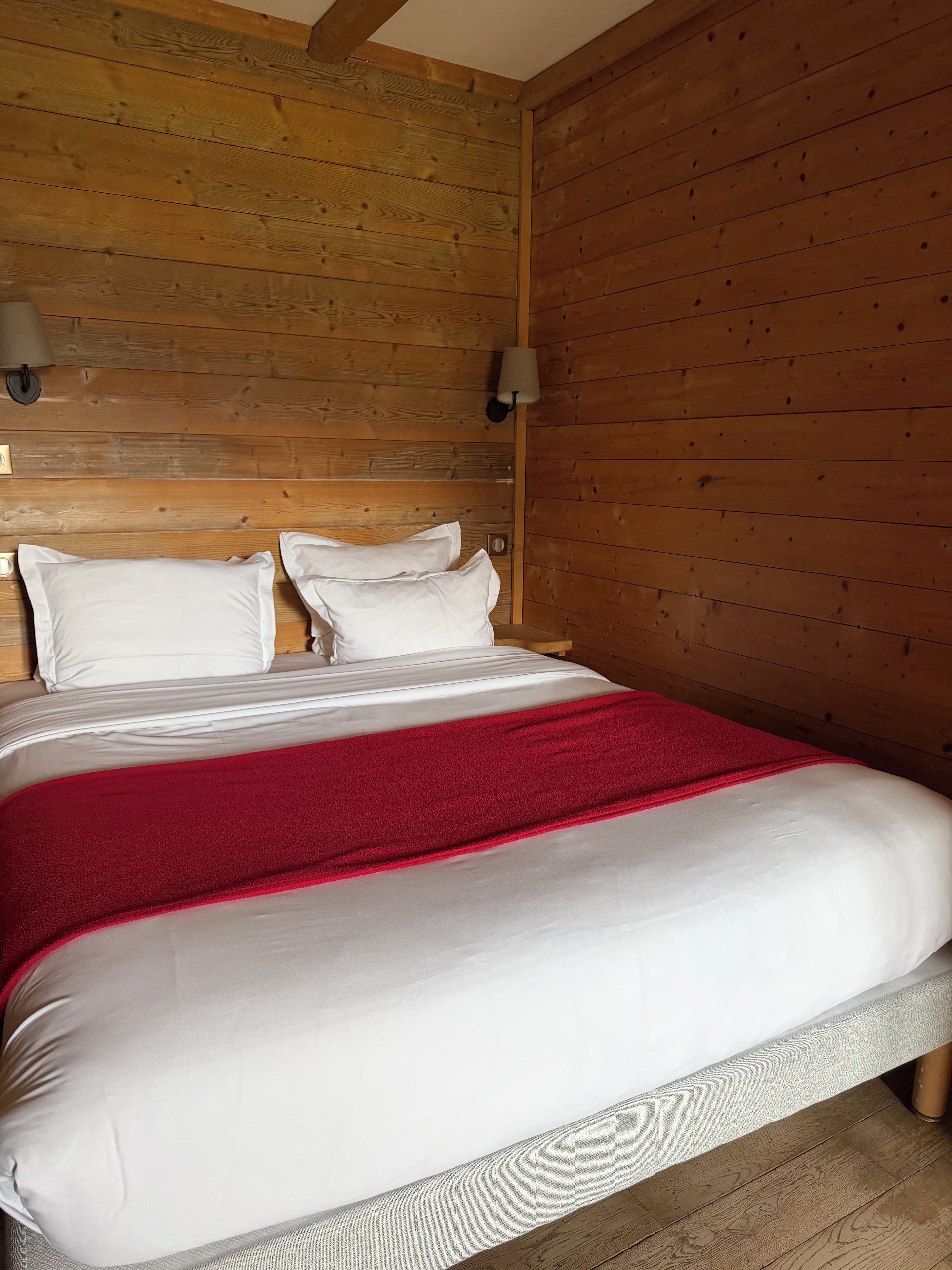 Ein Bett mit weißer Bettwäsche und einer roten Decke, Holzwände und einer Wandleuchte.