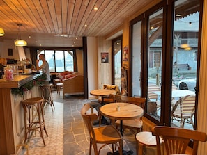 A cozy restaurant interior with wooden furniture, a bar area, and large windows offering a view of the outside. - Café-Hôtel Le Morillon (Morillon)