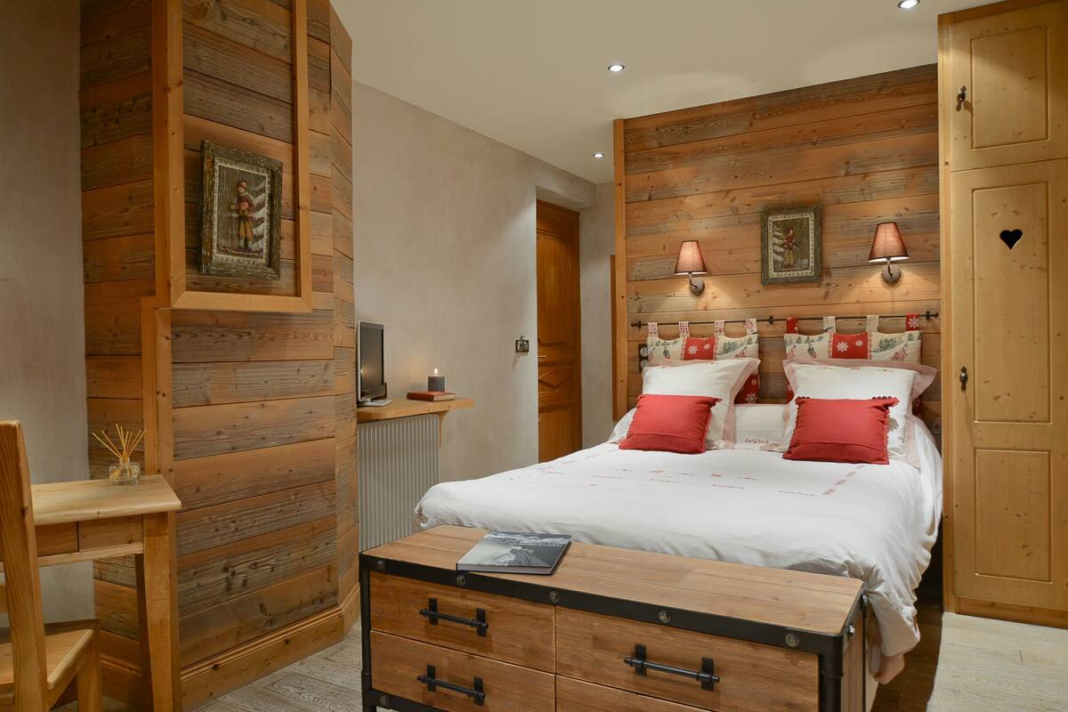 A bedroom with wooden panel walls, a bed with red and white bedding, a bedside table with a lamp, and a wooden chest of drawers.