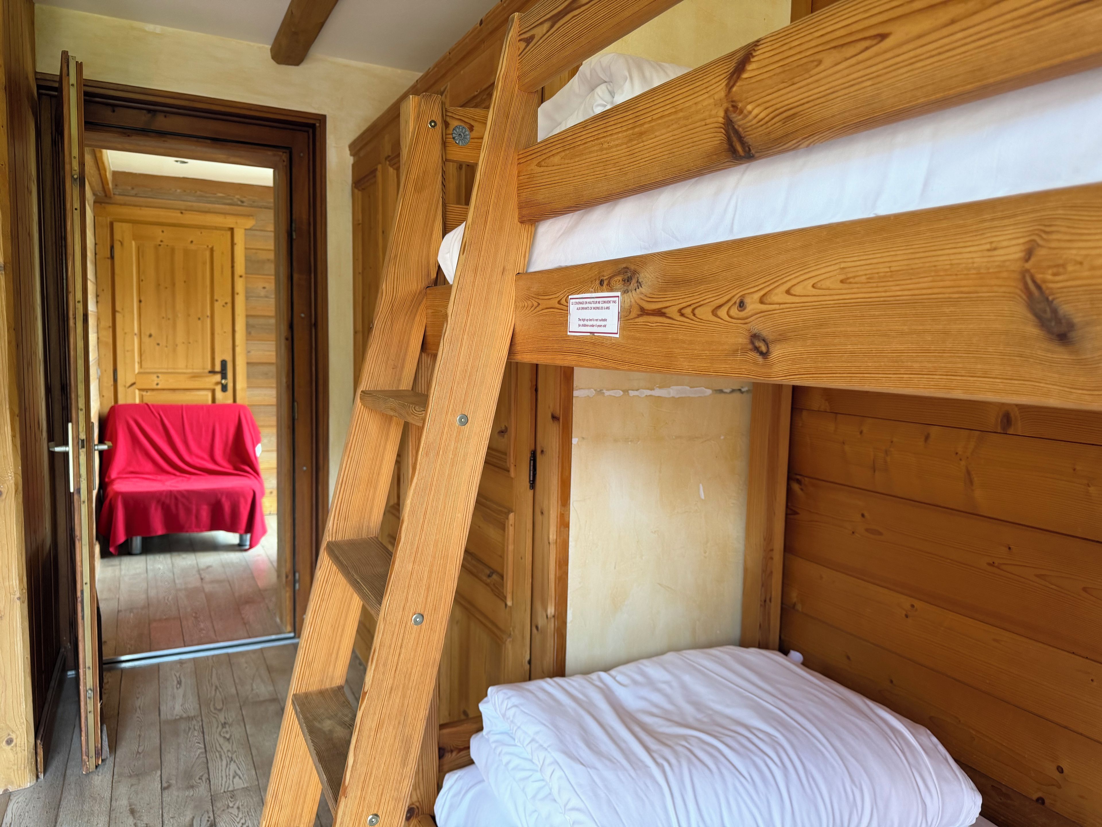 Une chambre avec un lit superposé en bois, une chaise recouverte de tissu rouge et une porte donnant sur une autre pièce.